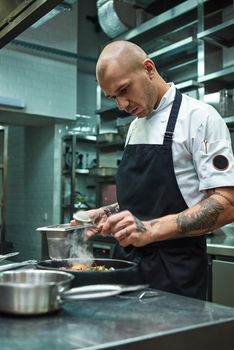 Need more spices. Vertical photo of restaurant chef with several tattoos adding a special spice to fresh cooked pasta Carbonara. Italian food