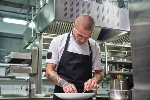 Finishing a dish. Attractive male chef with beautiful tattoos on his arms garnishing his dish on the plate in restaurant kitchen. Food decoration