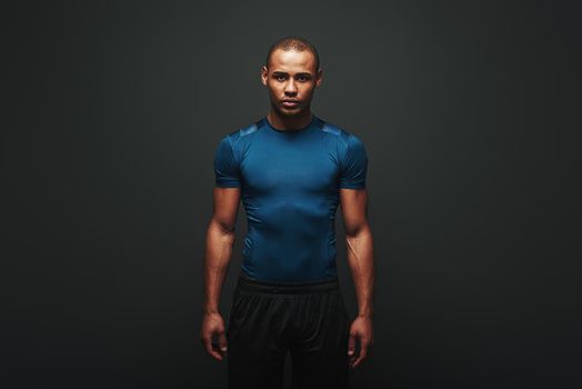 Portrait of handsome fitness man in blue T-shirt and black shorts standing isolated over dark background and looking at camera