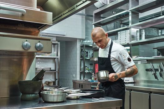 Sauce is ready. Handsome and confident chef in apron, with several tattoos on his arms holding a bowl with his famous sauce in a restaurant kitchen. Food concept