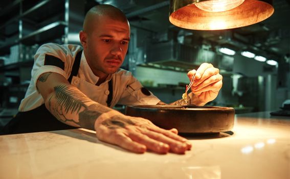 Ready to serve. Portrait of confident chef with tattoos on his arms decorating delicious chocolate cake under lamp light in a restaurant kitchen