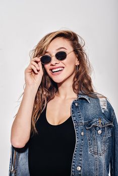 Girl with brown wavy hair. Stylish lady isolated over white background. Sensual gorgeous young woman in black bodysuit posing. She holds her sunglasses and smiling