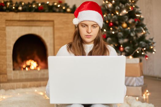 Happy woman looking in laptop in front of Christmas tree, lady wearing santa claus hat, looks at device screen, congratulating somebody via video call, posing in living room with x-mas decorations.
