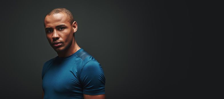 Portrait of handsome athlete in blue T-shirt and black shorts standing isolated over dark background and looking at camera. Long shot