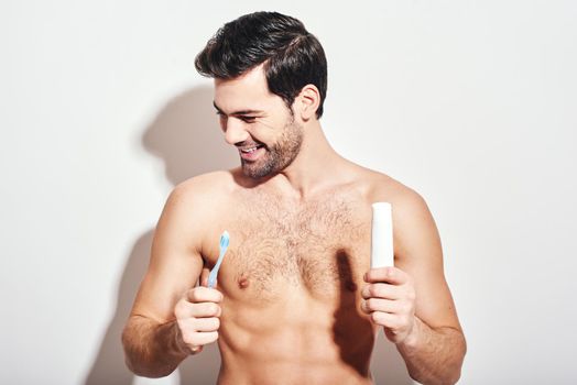 Handsome young man holding toothbrush and toothpaste in his hands while standing over white background. Idea of morning routine