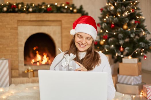Attractive Caucasian young smiling girl in santa hat having video call on her Laptop while sitting on floor at home near christmas tree and fireplace. Showing gift box to camera and smiling.