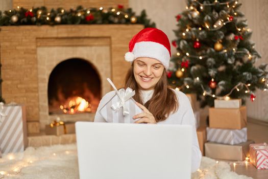 Happy smiling woman having video call via lap top, congratulated somebody with Christmas holidays and showing her gift to camera, lady wearing white casual sweater and santa hat.