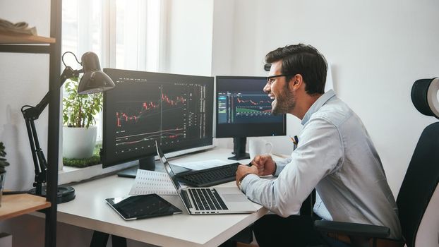 Great Happy young businessman or trader in formalwear looking at charts on computer screen in the office and smiling. Stock exchange. Financial trading concept. Investment concept