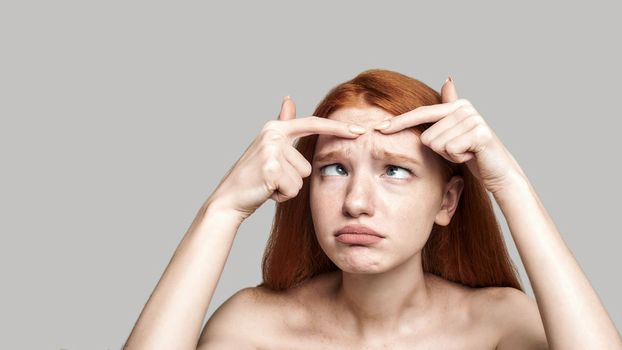 Studio shot of worried young redhead woman examining her face while standing against grey background. Skin care