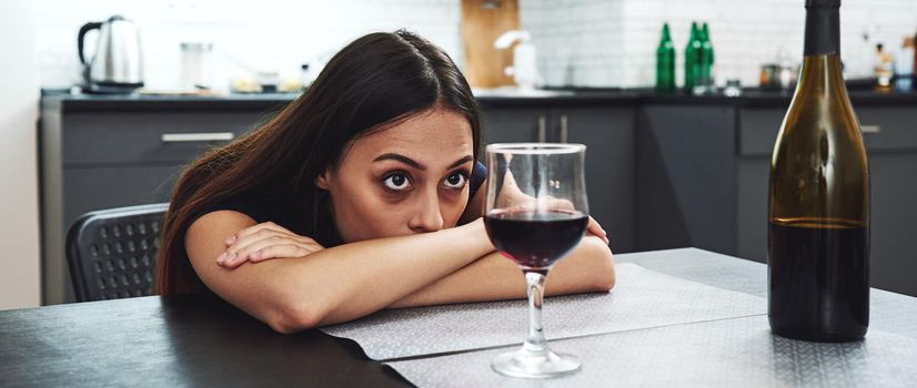 Dark-haired, sad and wasted alcoholic woman sitting at home, in the kitchen, leaning over the table, looking at bottle and glass of red wine on the table, completely drunk, looking depressed, lonely and suffering hangover in alcoholism and alcohol abuse