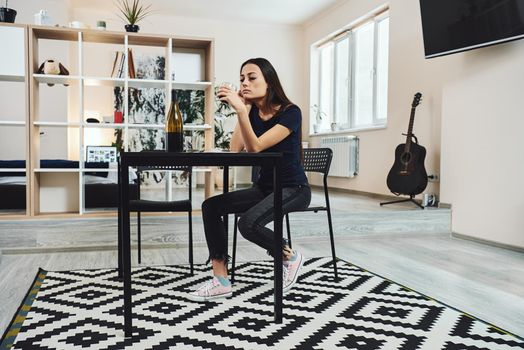 Depressed, divorced woman sitting alone in kitchen at home and holding a glass of red wine because of problems at work and troubles in relationships. Social and life problems. Full-length shot.