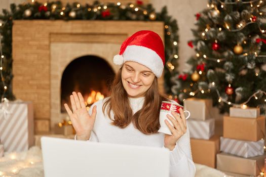 Close up smiling European woman using laptop, looking at screen, waving hand, greeting, chatting online with somebody, making video call on Christmas tree and fireplace background at home.