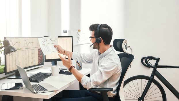 Growing chart. Side view of happy young bearded trader in headset holding financial report, talking with client and smiling while sitting in front of monitor screens in the office. Business concept. Trade concept. Call center