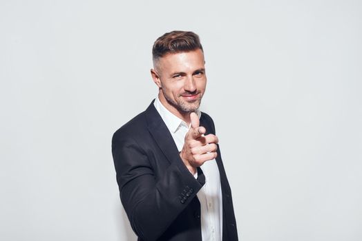 Studio shot of cheerful bearded businessman in classic suit gesturing and smiling while standing against grey background. Men beauty. Business look. Studio shot