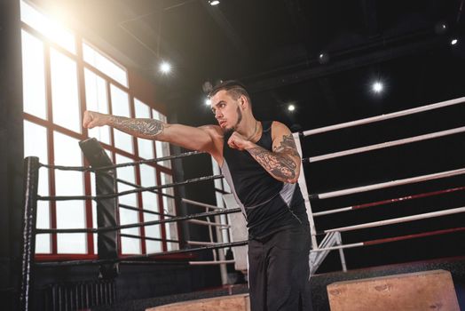 Muscular athlete in sports clothing practicing right straight punch in boxing gym. Strong young man doing shadows-boxing