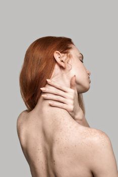 Studio shot of back view of young and beautiful redhead woman touching her neck while standing against grey background