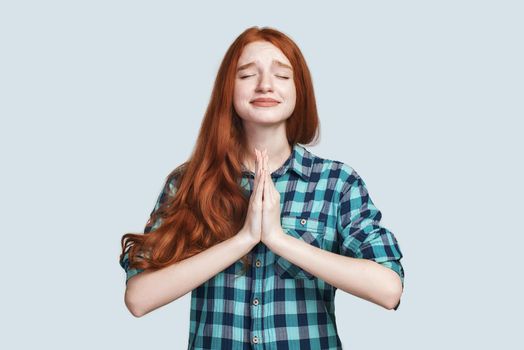 Studio shot of redhead girl in checkered shirt worrying, holding hands in pray, apologizing for bad behaviour or mistake, standing over grey background with closed eyes