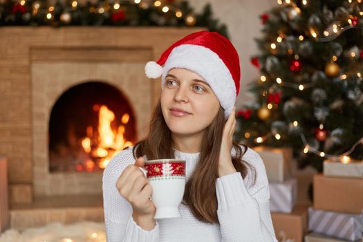 Young pensive woman in Santa hat by Christmas tree and fireplace at home, looking dreamily aside, holding cup with coffee or tea, wearing white warm sweater.