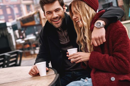 Drink coffee together. Young couple are speaking while drinking hot beverage, tea or coffee outdoors. They are sitting on the bench, young man huggs his girlfriend