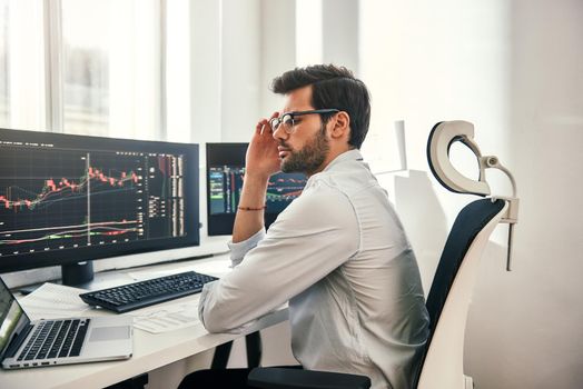 Full concentration. Young and smart businessman in formal wear is adjusting his eyeglasses and using laptop while while sitting in his office in front of computer screens with trading charts. Stock exchange. Trade concept. Investment concept