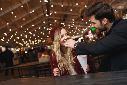 One french fries for two. Young couple are eating french fries at the street food market. Cold season. Close-up photo of couple eating from one package