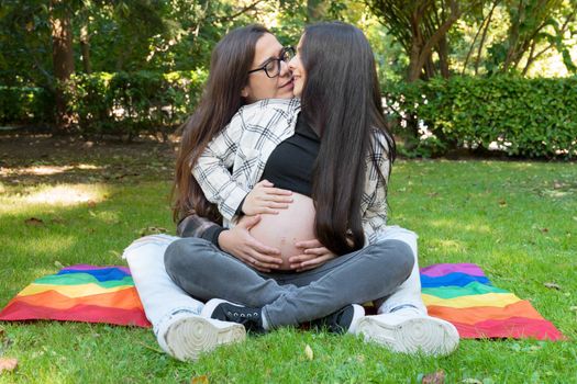Portrait of affectionate pregnant lesbian couple with rainbow flag, relaxed at the park. Two happy girlfriends. Homosexual relationship. LGBT Community Pride. High quality 4k footage