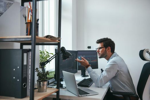 Really Side view of surprised young male stock market broker analyzing a graphs on computer and gesturing while sitting in his modern office. Stock exchange. Trade concept. Investment concept