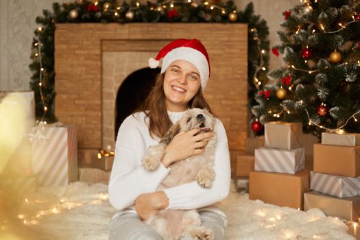 Happy family in red hat and white casual sweater with dog sitting near christmas tree, present boxes and fireplace, cheerful girl with her pet, puppy biting owner's finger.