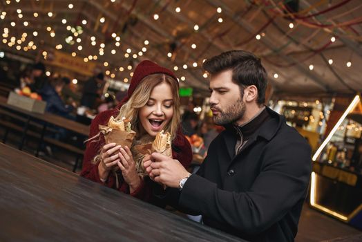 Give me a bite . Young couple are eating sandwiches at the street food market. Cold season. Close-up photo of couple are eating at food cort