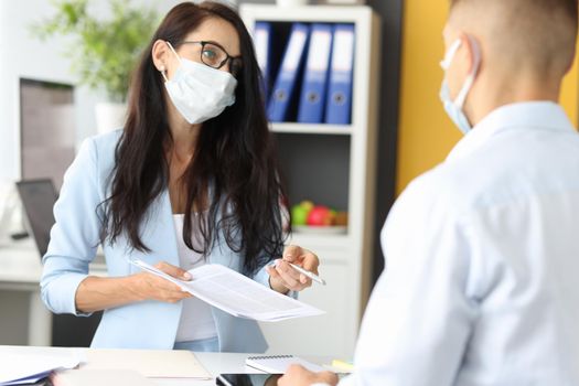 A woman in a protective mask at the clinic's reception speaks to a man. Filling out a questionnaire for visiting a doctor