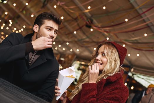 Couple are eating outdoors . Young couple are eating french fries at the street food market. Cold season. Close-up photo of couple eating from one package