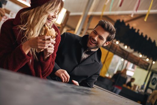 Dont touch my food . Young couple are eating sandwiches at the street food market and having fun together. Cold season. Woman take away her sandwich from a man for fun