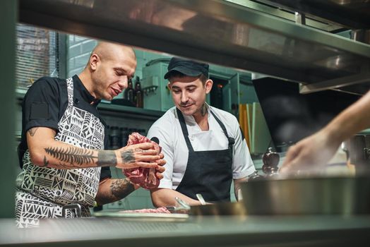 How to choose the right meat Professional chef in apron and with tattoos on his arms showing a red meat to his assistant while standing in a restaurant kitchen.