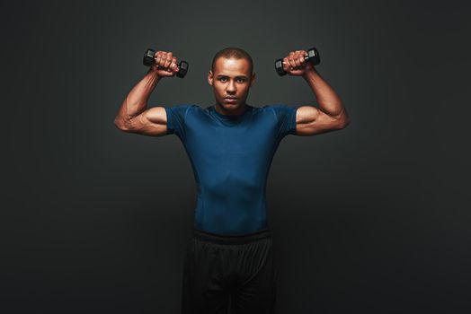 Athletic young sportsman exercising with dumbbells and looking at camera isolated on dark background. He demonstrates his perfect muscles