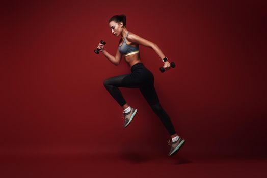 Full length portrait of a healthy muscular sportswoman jumping isolated over red background with dumbbells in her hands. Dynamic movement. Side view