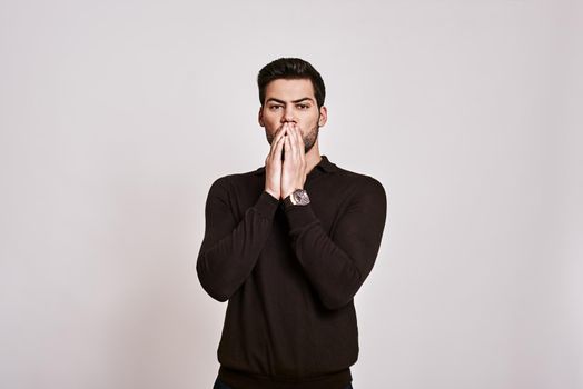 Portrait of surprised, shocked man with stubble and widened eyes in brown jumper touching his face with two hands isolated on grey background