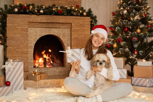 Cute happy female sitting on floor with crossed legs, showing to camera her Christmas present box and hugging her dog, looks smiling directly at camera, wearing casual attire and red hat.