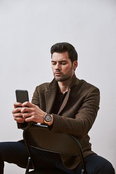 Isolated studio shot of handsome young male corporate worker with bristle and stylish haircut looking at his phone while sitting on a chair isolated over white background.