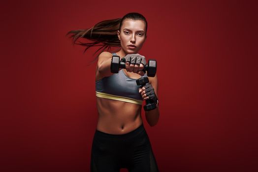 Portrait of a confident young fitness woman doing exercises with dumbbells isolated over red background. Dynamic movement. Studio shot