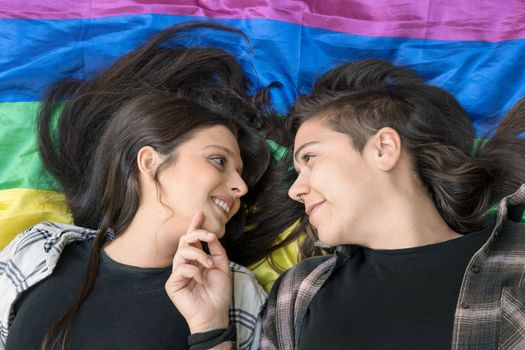 Lesbian couple wearing a lgbt rainbow flag lying on the floor. Affectionate moment between two women under a lgbt flag. High quality photo.