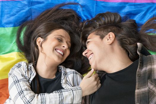 Lesbian couple wearing a lgbt rainbow flag lying on the floor. Affectionate moment between two women under a lgbt flag. High quality photo.