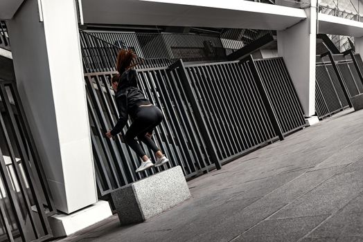 Young woman doing leg exercises at stoned step platform outside with city on background. Beautiful cauceuropean woman jumping over big step, squat exercise. Fitness and health concepts. Rear view