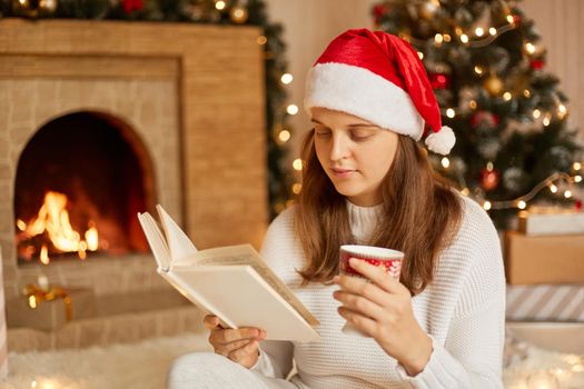 Woman resting with cup of hot drink and book near fireplace and Christmas tree, looking concentrated on pages, spending leisure time, wearing festive hat and white knitted sweater.