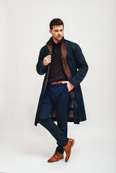 Full-length portrait of stylish dark-haired man wearing blue trousers, brown jacket and blue coat standing, holding one hand in trousers pocket and looking away isolated over white background