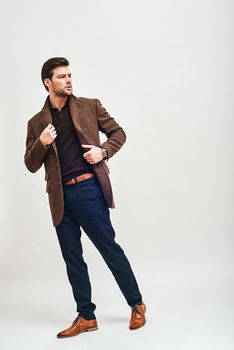 Full-length portrait of stylish dark-haired man wearing blue trousers and brown jacket, standing and looking away isolated over white background