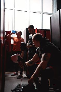 Muscular athletes boxers in sports clothing preparing colored boxing gloves for training in dark gym locker room