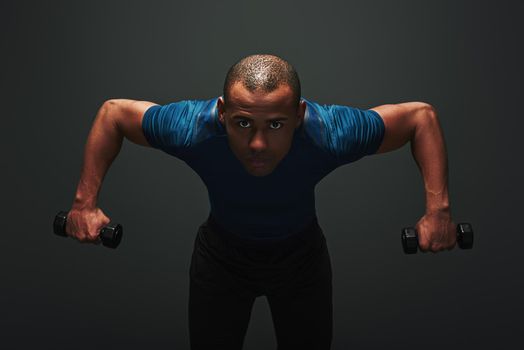 Athletic young sportsman exercising with dumbbells and looking at camera isolated on dark background