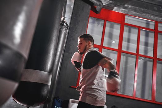 Handsome young muscular sportsman in sports clothing boxing on heavy punch bag. Hard training before big fight in colored boxing gym