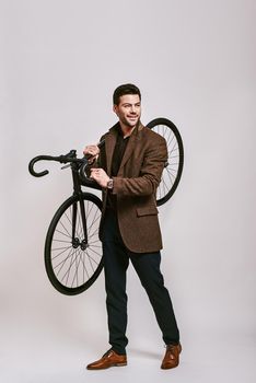 Full-length portrait of stylish dark-haired man wearing blue trousers, brown jacket holding bicycle in his hands, smiling and looking away isolated over grey background
