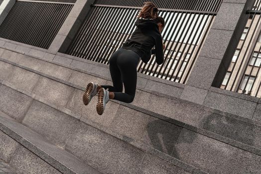 Young woman doing leg exercises at stone stairs outside with city on background. Beautiful cauceuropean woman jumping over big step, squat exercise. Fitness and health concepts. Rear view